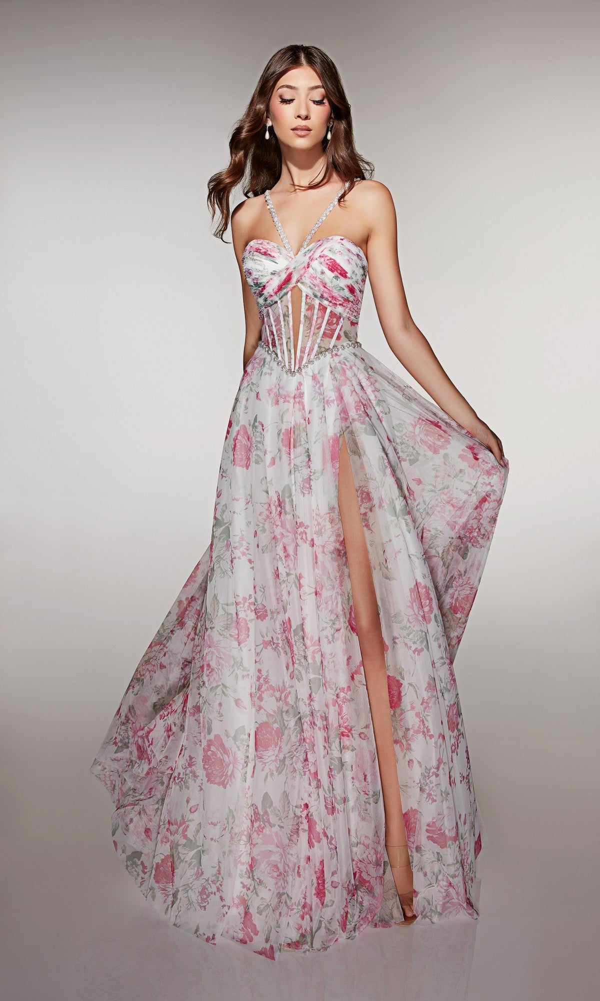 A woman wears the Alyce 61994 long prom dress featuring pink floral print, a sheer corset bodice, spaghetti straps, and a high leg slit, standing against a plain background.